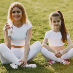 Smiling mother and daughter wearing matching outfits sitting on grass in a park.